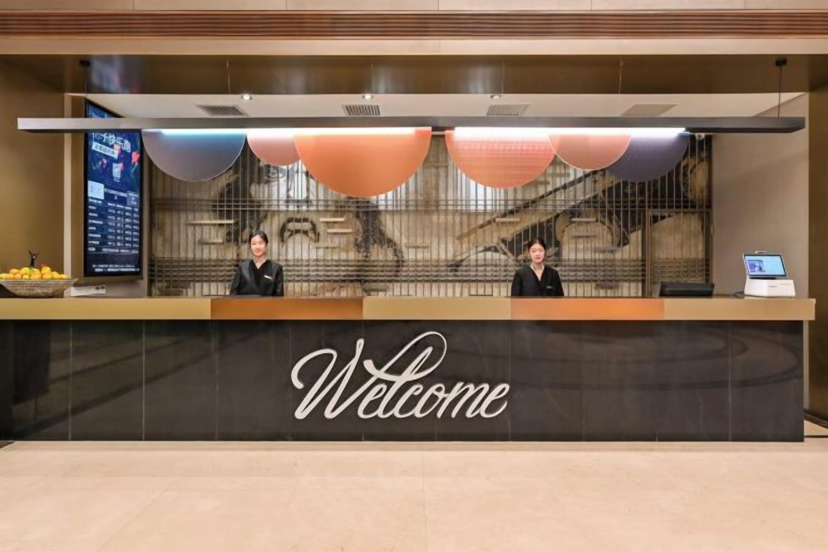 Una recepción con un cartel de 'Welcome', dos empleados, una pantalla digital y una exposición de frutas.