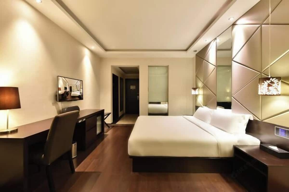 A modern hotel room with a large bed, a desk, and a chair. There is a mirror on the wall and a door leading to another room.