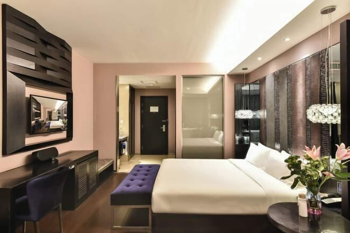 A modern hotel room with a large bed, a desk, a mirror, and a bathroom visible through a glass door.
