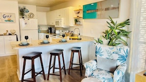 Una cocina moderna con armarios blancos, una isla central con taburetes, una planta grande en maceta y un sillón azul y blanco con estampado floral.