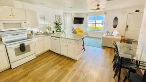 Una cocina moderna con armarios blancos, microondas y horno. Hay un comedor con mesa y sillas de vidrio.