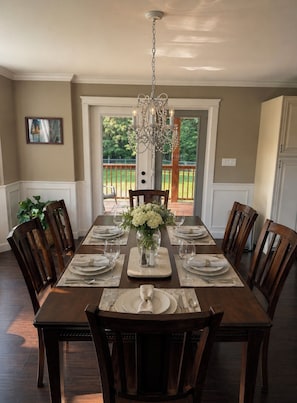 A dining room with a long table set for six, surrounded by wooden chairs, a chandelier, and a view of a deck. - Caribou Lodge ~ Entire private home with hot tub! (Terrace)