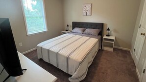 Un dormitorio con una cama grande, dos mesitas de noche, un tocador, un televisor, una ventana con persianas y una obra de arte colgada en la pared.