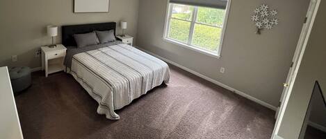 A bedroom with a large bed, a nightstand, a lamp, a window, and a decorative wall piece.