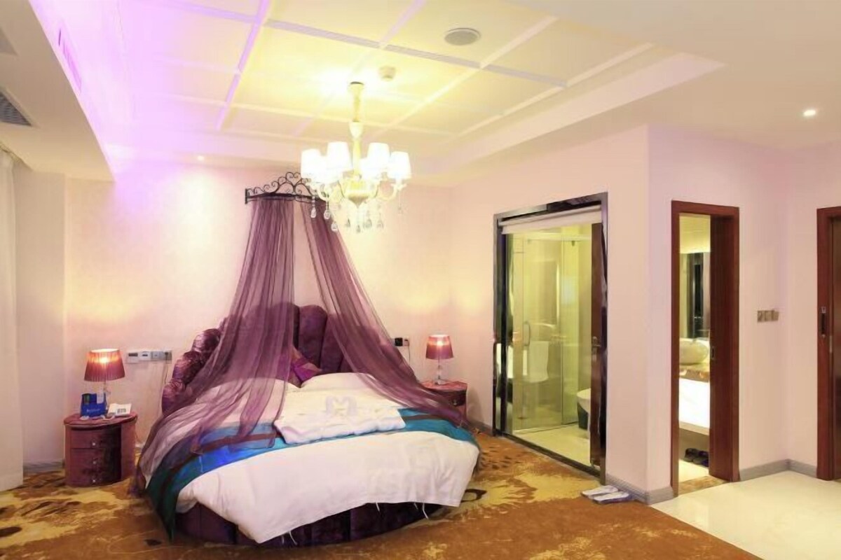 A bedroom with a canopy bed, a chandelier, a glass door leading to a bathroom, and a carpeted floor.