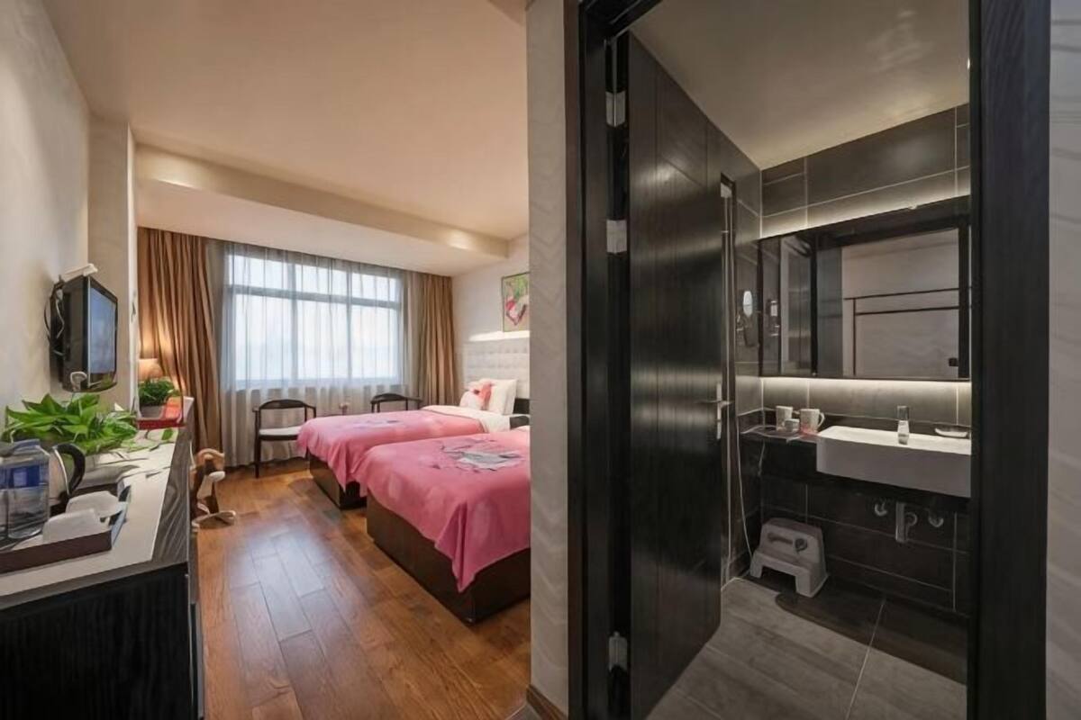 A hotel room with a desk, two beds, a TV, a desk lamp, a chair, a small table with a plant, and a bathroom with a sink and mirror.