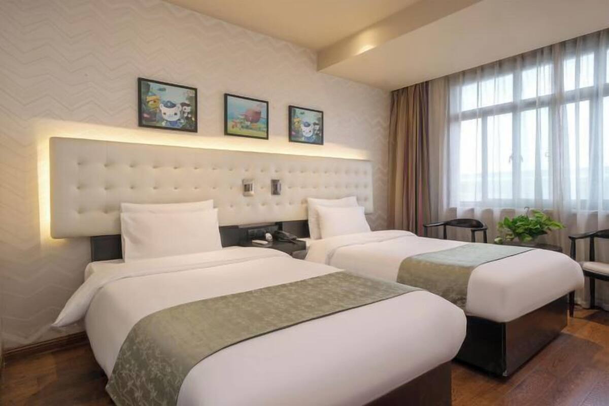 A hotel room with two beds, a large padded headboard, a desk with a chair, and a large window with curtains.
