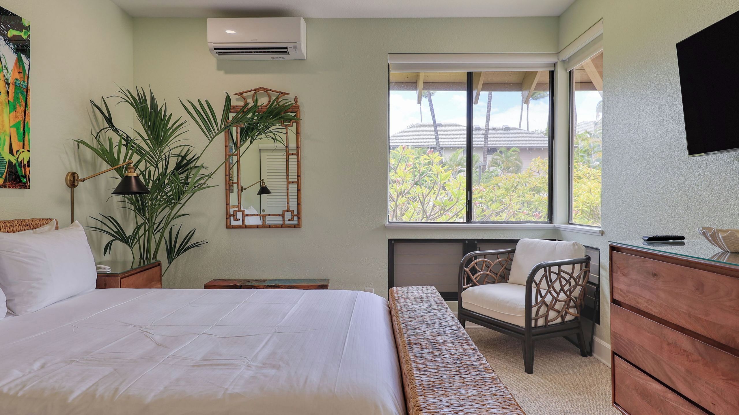 Quarto com cama, mesa de cabeceira, plantas em vasos, uma cadeira, cômoda, janela com vista e televisão fixada na parede.