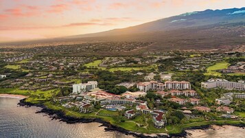 An aerial view of a coastal resort area with multiple buildings, including hotels and residential structures, surrounded by greenery and a beach.