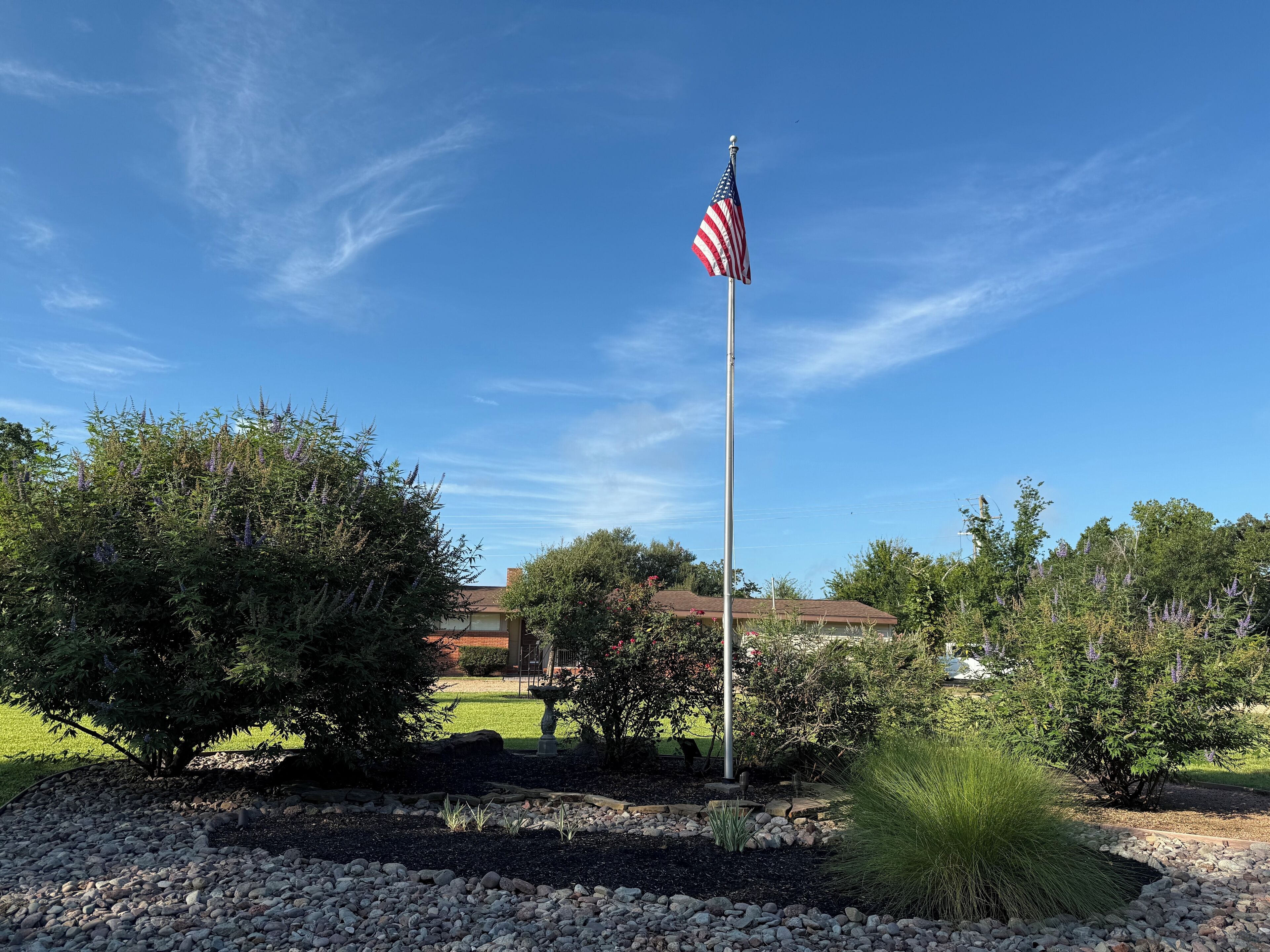 A flagpole with an American flag, surrounded by landscaped garden beds and trees.