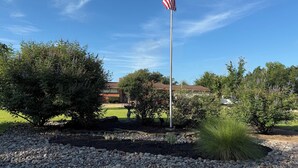 A flagpole with an American flag, surrounded by landscaped garden beds and trees. - NEAR CAMPUS! Choice KING or QUEEN BEDS w fresh interior paint & great parking! (College Station)