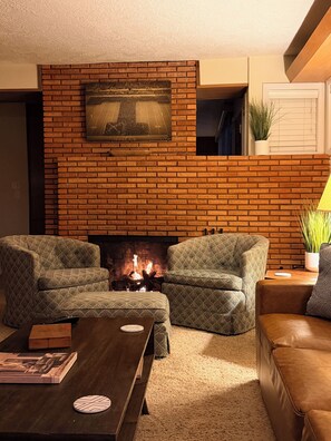 A living room with a brick fireplace, two patterned armchairs, a brown leather sofa, a coffee table, and a television mounted on the wall.