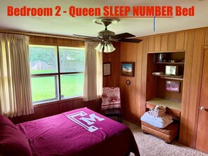 Ein Schlafzimmer mit einem Queensize-Bett, Holzpaneelen, einem Deckenventilator, einem Fenster mit Vorhängen und einem Bücherregal mit gerahmten Bildern.