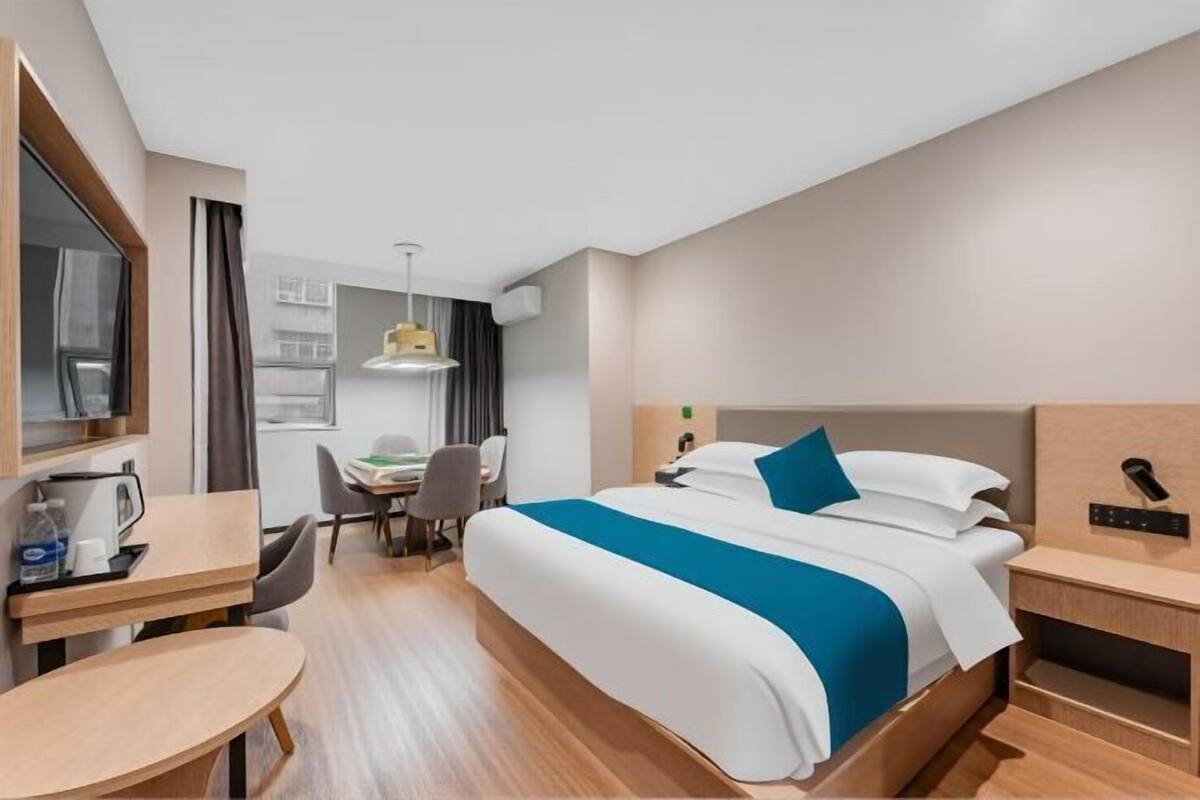 A hotel room with a bed, a desk, a chair, a small table, and a desk lamp.