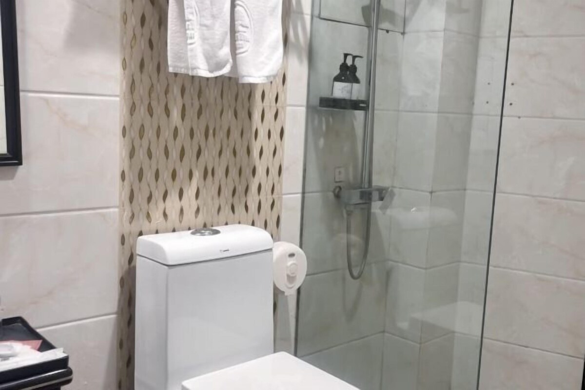 Ein modernes Badezimmer mit Duschkabine aus Glas, einer weißen Toilette und einem waschraumtypischen, gemusterten Duschvorhang.