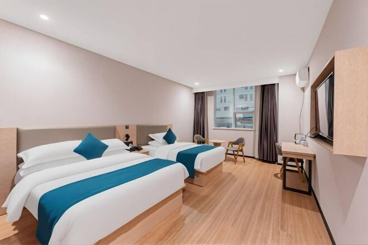 A hotel room with two beds, a desk, a chair, a window with curtains, and a view of buildings outside.