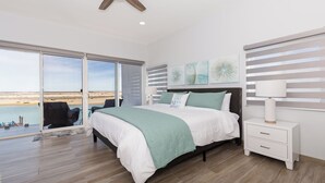 Un dormitorio con cama, mesita de noche, lámpara y vista a la playa a través de una puerta corrediza.