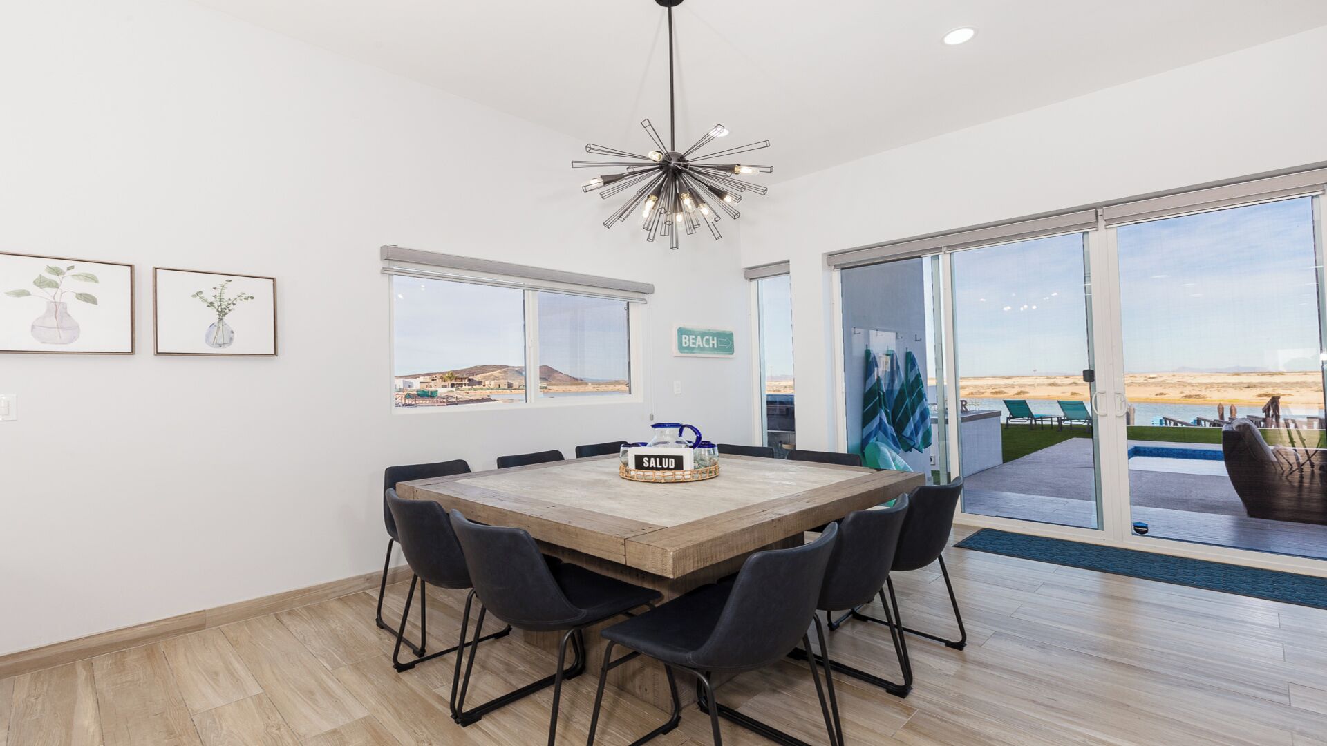 Un comedor amplio con una mesa de madera y sillas negras, ventanas grandes con vistas a la playa, y un cartel que indica el nombre de la habitación.