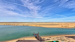 Paisaje desértico con cielo azul despejado, una masa de agua y un muelle de madera que se adentra en el agua.