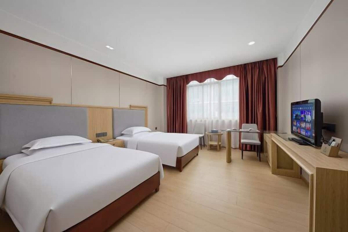 A hotel room with two beds, a desk with a TV, and a window with curtains.