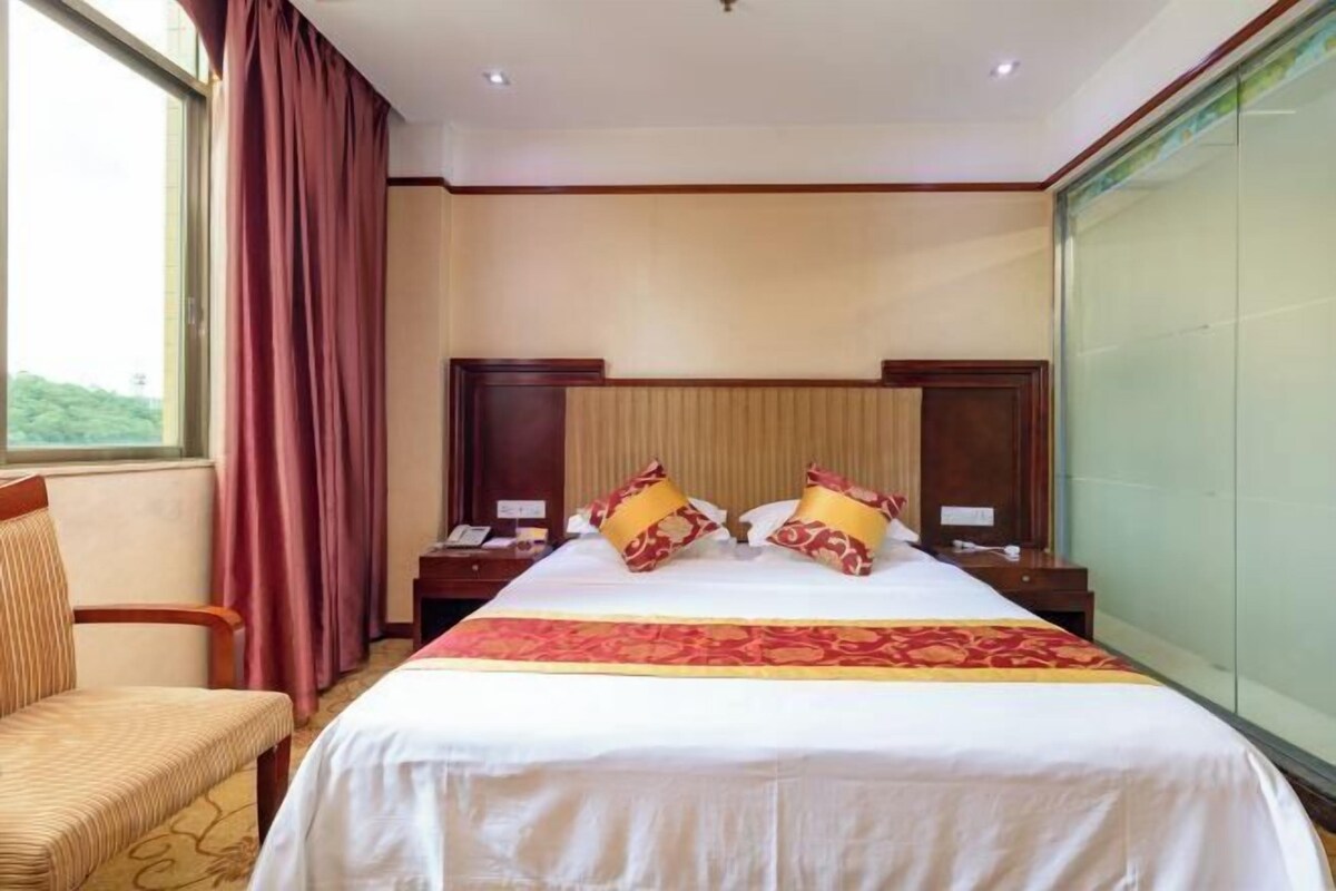 Una habitación de hotel con una cama grande, dos mesitas de noche, una silla y un ventanal con cortinas.