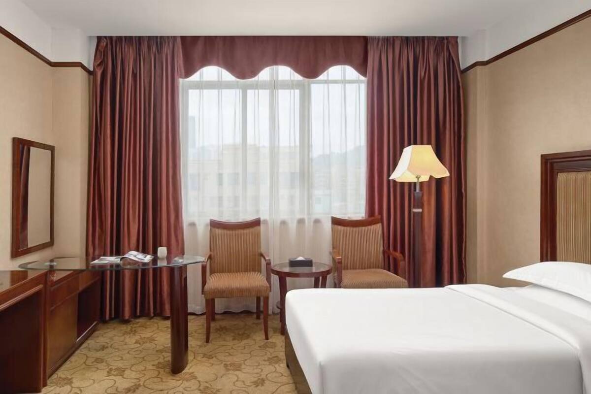 A hotel room with a bed, two chairs, a small table, a lamp, and a large window with curtains.