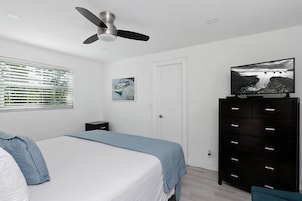 Quarto com uma cama, um ventilador de teto, uma TV sobre um criado-mudo, uma janela com persianas e um quadro na parede.