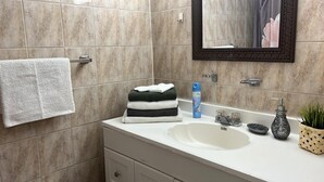 A bathroom with beige tiles, a white sink, a mirror, a towel rack, a stack of folded towels, a soap dispenser, a small bowl, and a potted plant. - Comfortable, Within Walking Distance To The Beaches 2217
 (Hallandale Beach)