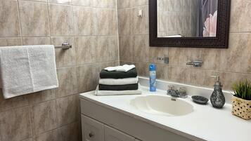 Une salle de bain avec des carreaux de couleur beige, un lavabo blanc, un miroir, un porte-serviettes, une pile de serviettes pliées, un distributeur de savon, un petit bol et une plante en pot.