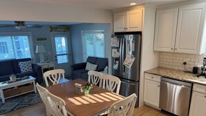 A modern kitchen with white cabinetry, a stainless steel refrigerator, and a dishwasher. There is a dining table with white chairs and a blue sofa in the living area. - Charming 3-bedroom house in beautiful Cape May, NJ (Cape May)