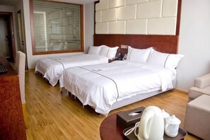 Ett hotellrum med två sängar, ett trägolv, en sänggavel med en ljus panel, ett litet bord med en telefon och en kopp, och ett fönster med persienner.