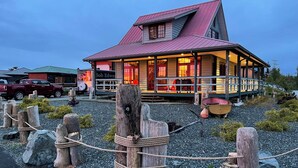 Un chalet avec un toit rouge et une terrasse en bois, entouré d’un chemin de gravier et de poteaux en bois.