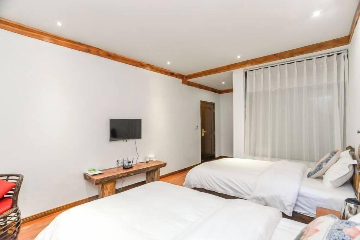 Habitación de hotel con dos camas, un banco de madera, un televisor de pantalla plana y un ventanal con cortinas.