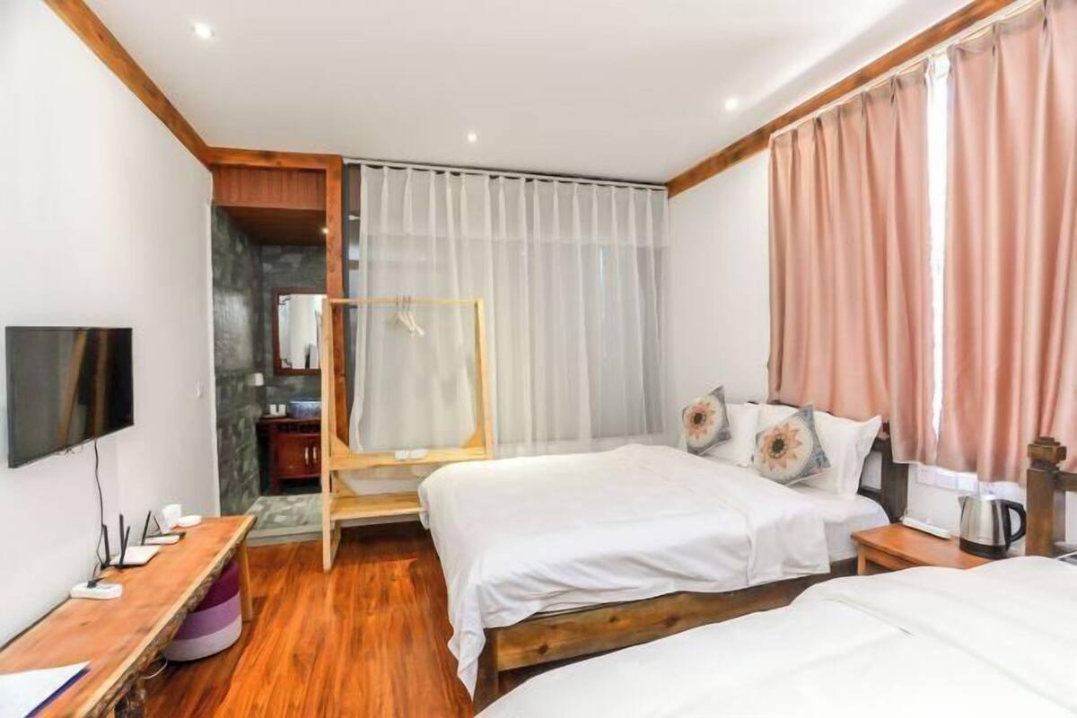 Habitación de hotel con dos camas, un escritorio de madera, televisor, zona de ducha y un amplio ventanal con cortinas.