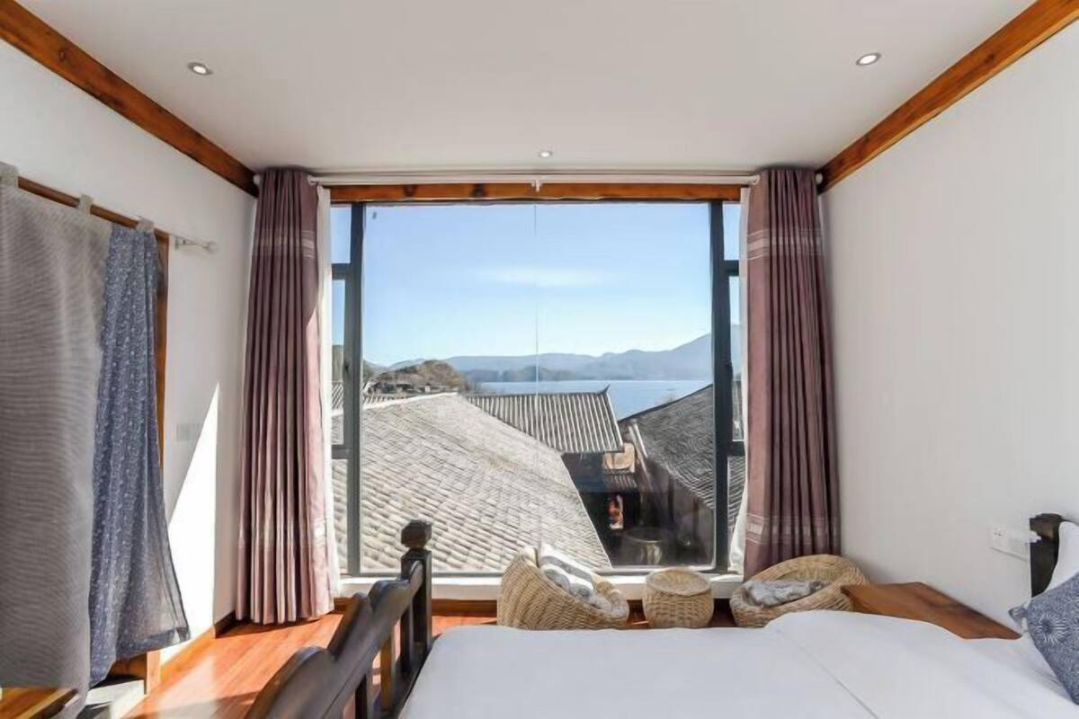 Un dormitorio con un ventanal grande, vigas de madera, una cama y vistas a un edificio tradicional y montañas.