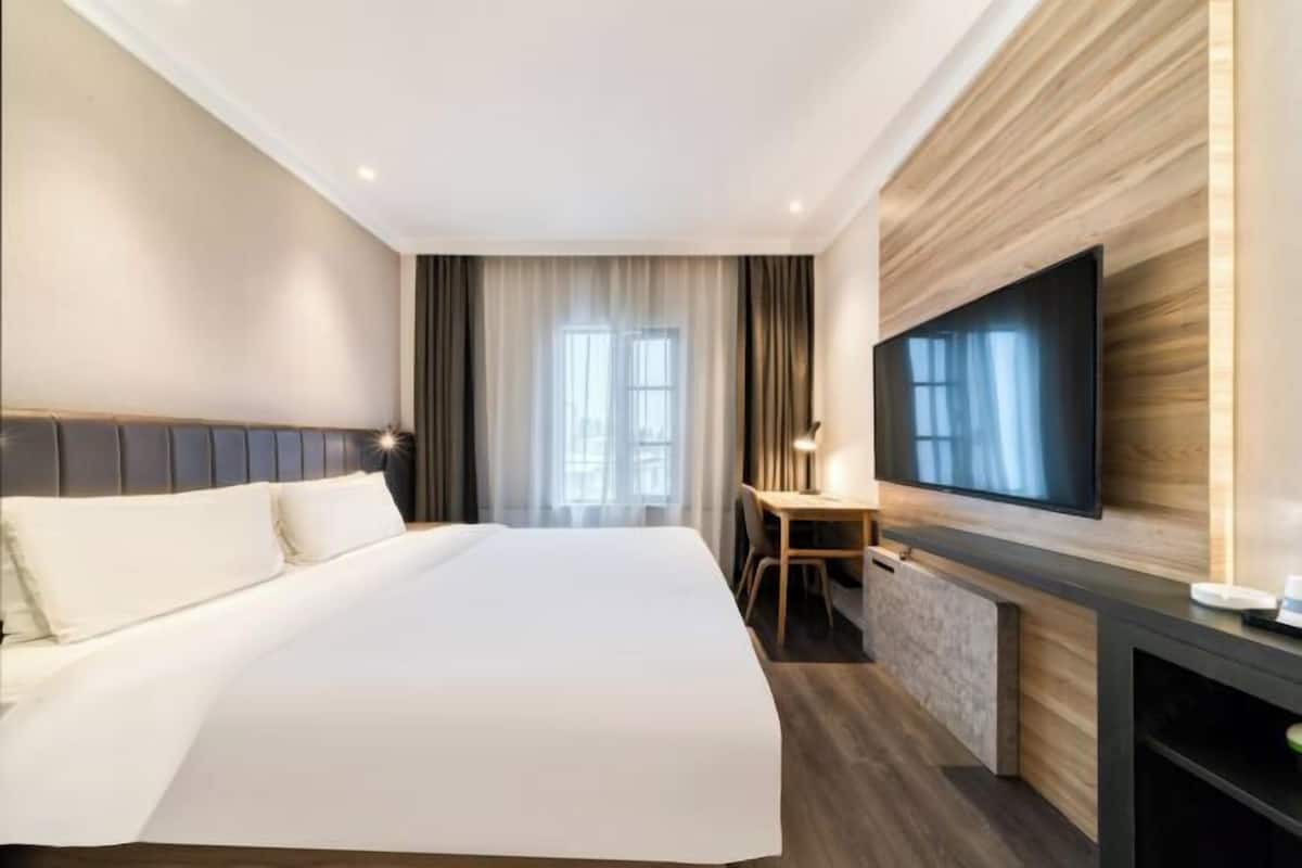 A hotel room with a large bed, a TV mounted on the wall, a desk, and a window with curtains.