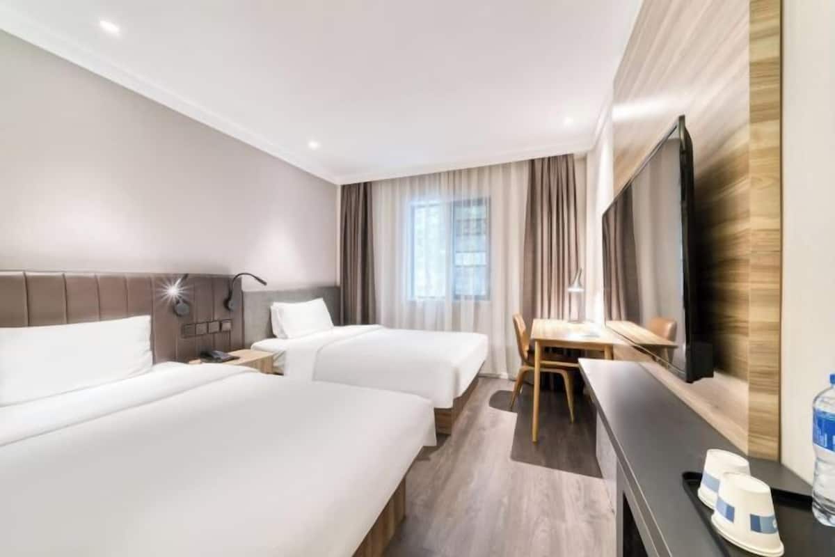 A hotel room with two beds, a desk, a chair, a TV, and a large window with curtains.