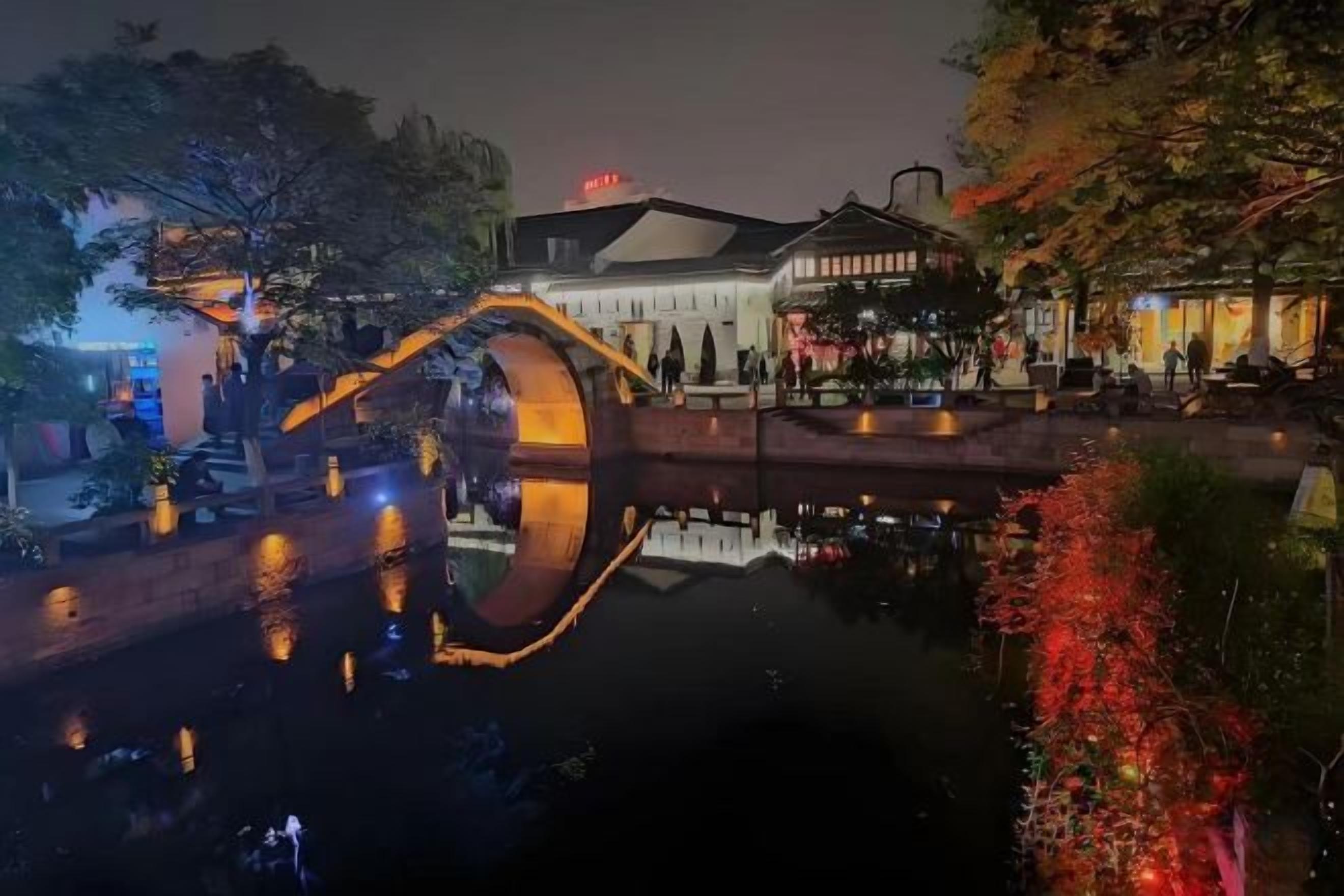 A traditional building with a large arch bridge over a body of water, surrounded by trees and illuminated by ambient lighting.