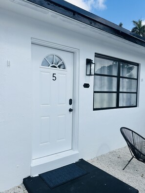 A white house with a number 5 door, a window, and a black chair outside. - Wilton Manors Guesthouse (Wilton Manors)