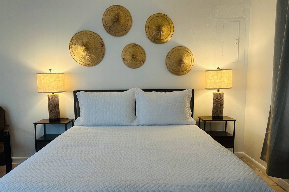 Una cama bien hecha con un edredón blanco acolchado, flanqueada por dos mesitas de noche con lámparas y un objeto decorativo en la pared.