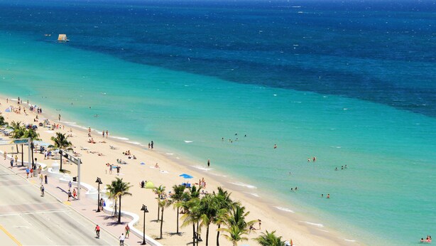 Een strand met helder turquoise water, palmbomen en een brede boulevard.