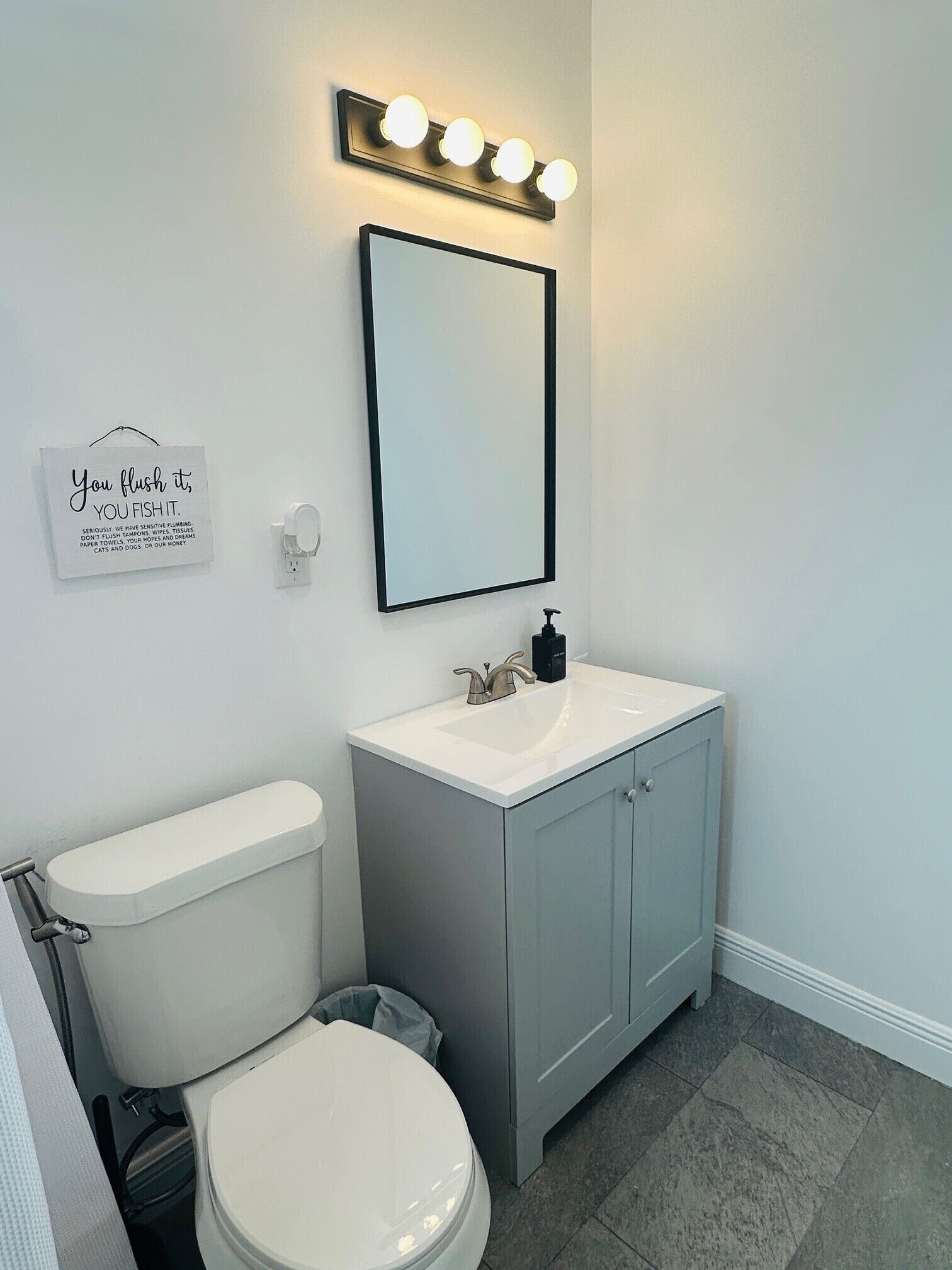 A bathroom with a toilet, sink, cabinet, and a mirror with lights. There's a sign on the wall.