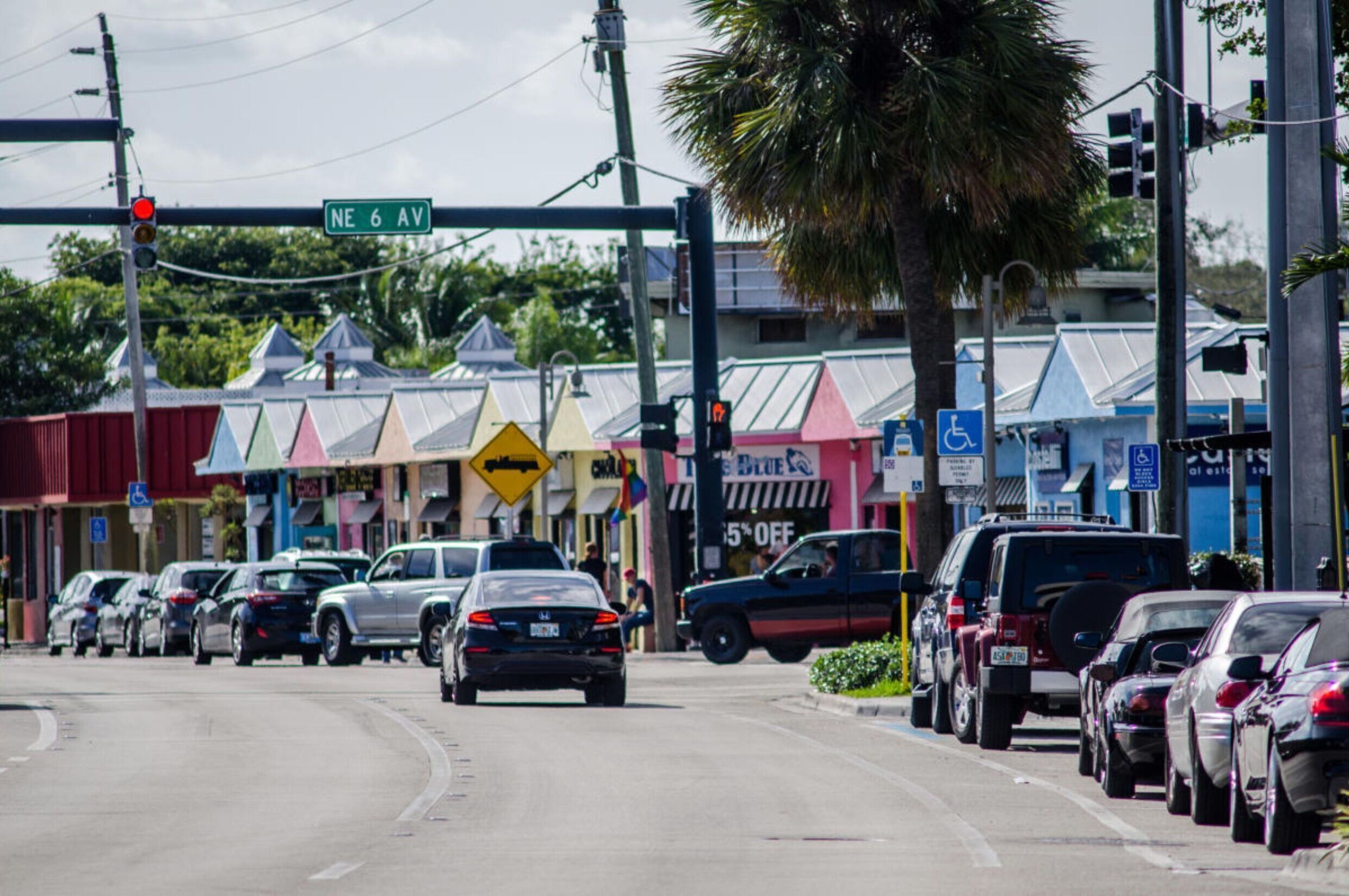 A street view with a row of parked cars, a traffic light, and a street sign for NE 6 AV.