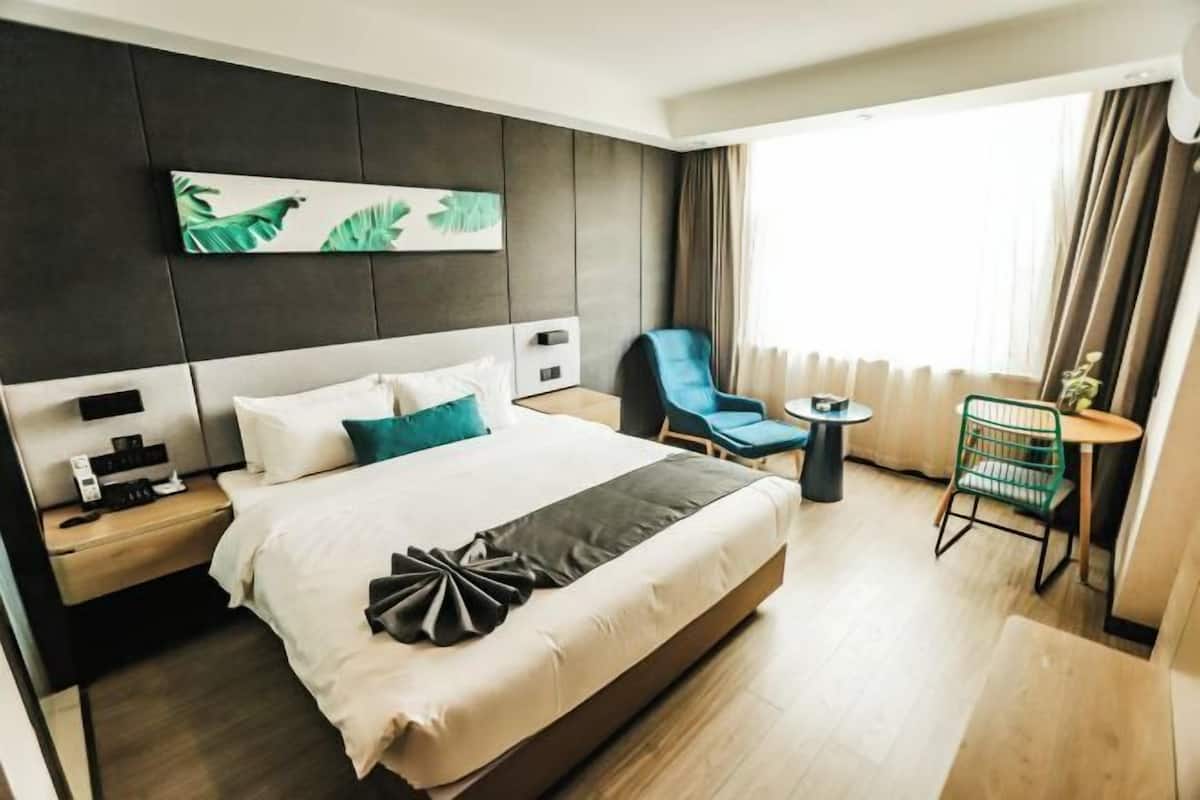 A modern hotel room with a large bed, a bedside table with a phone and charger, a chair, a small table, and a window with curtains.