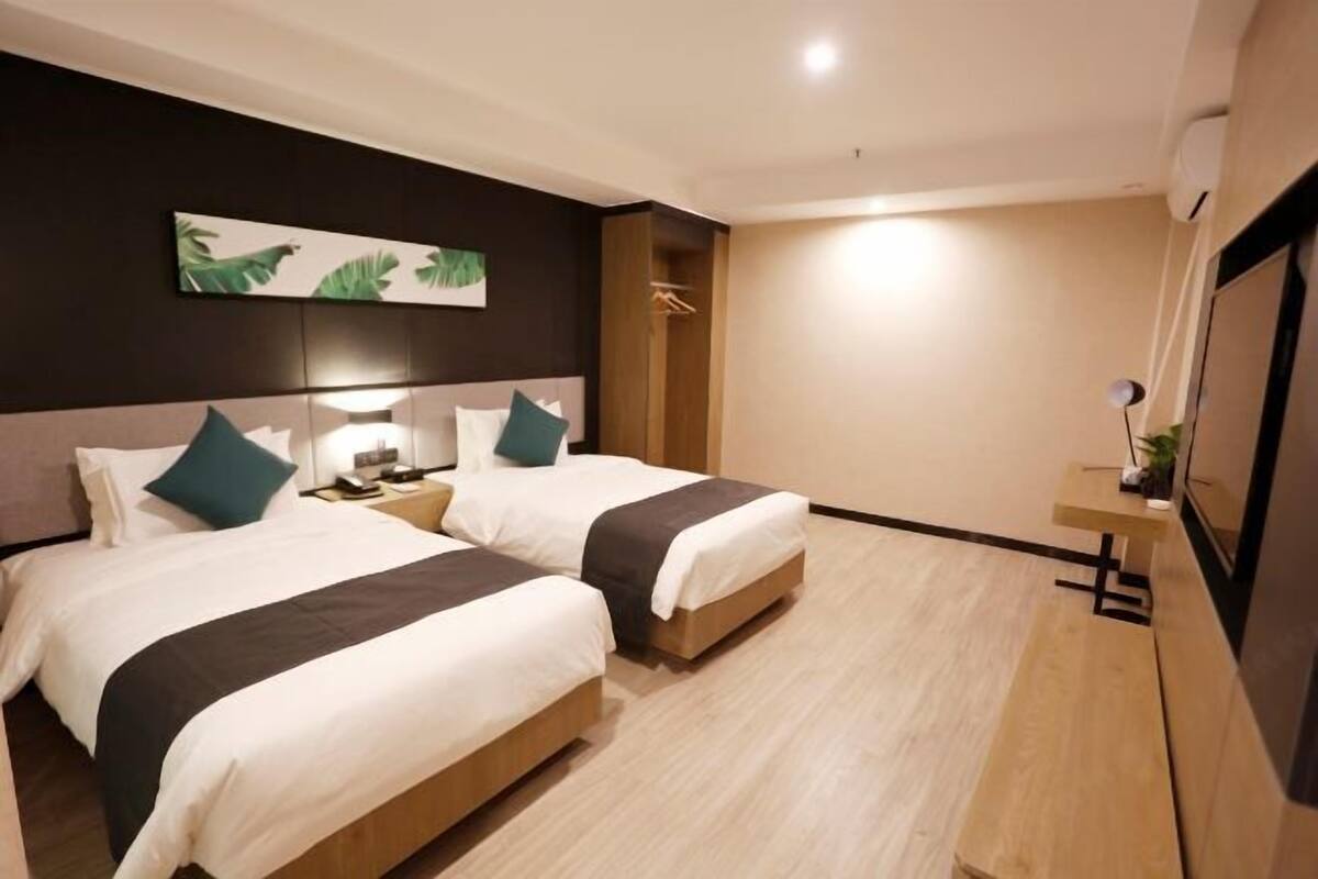 A hotel room with two beds, a nightstand, a bedside table, a wall-mounted TV, and a wooden bench.