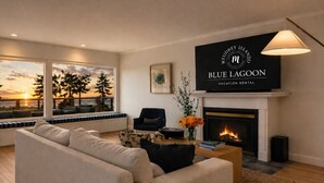 A modern living room with a sofa, a fireplace, a television displaying 'Blue Lagoon Vacation Rental,' and a view of trees through large windows.