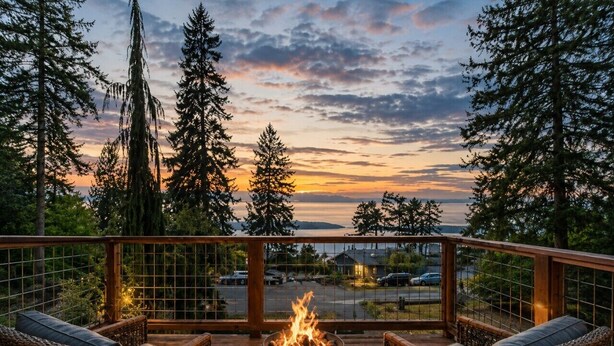 A deck with wicker furniture and a central fire feature, overlooking a parking area and trees.