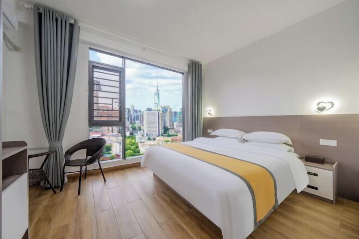 A hotel room with a bed, a desk, a chair, a nightstand, and a view of a cityscape through a large window.