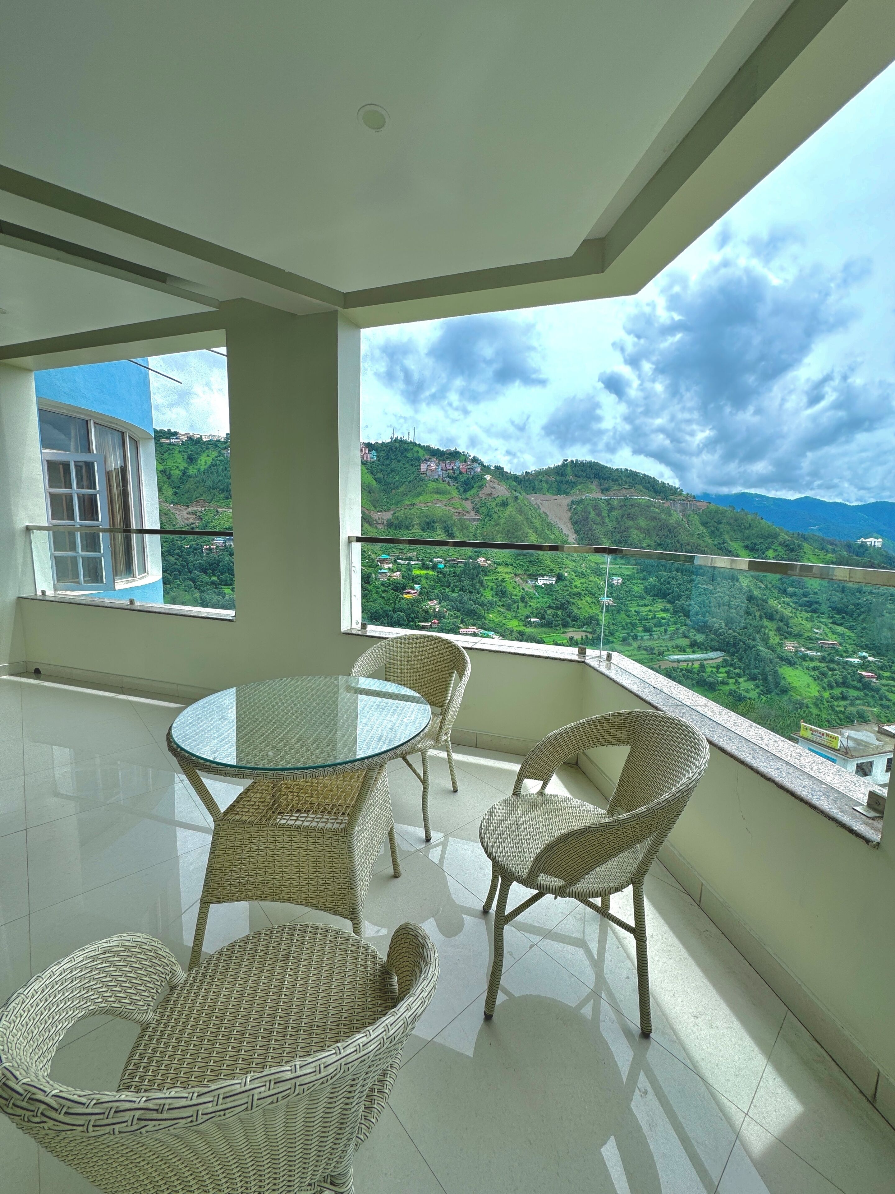 A balcony with a glass table and wicker chairs, overlooking a green valley with buildings on hills.
