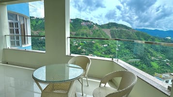 A balcony with a glass table and wicker chairs, overlooking a green valley with buildings on hills.